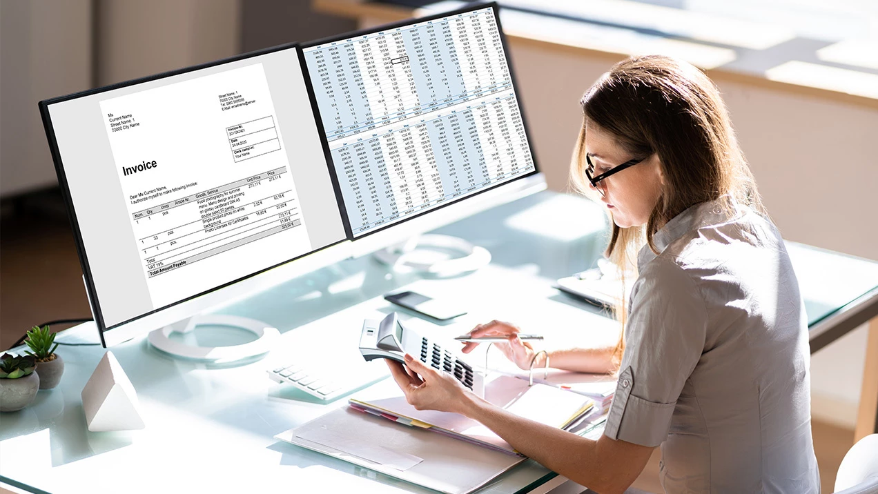Person working at a desk with dual monitors displaying an invoice and spreadsheet.